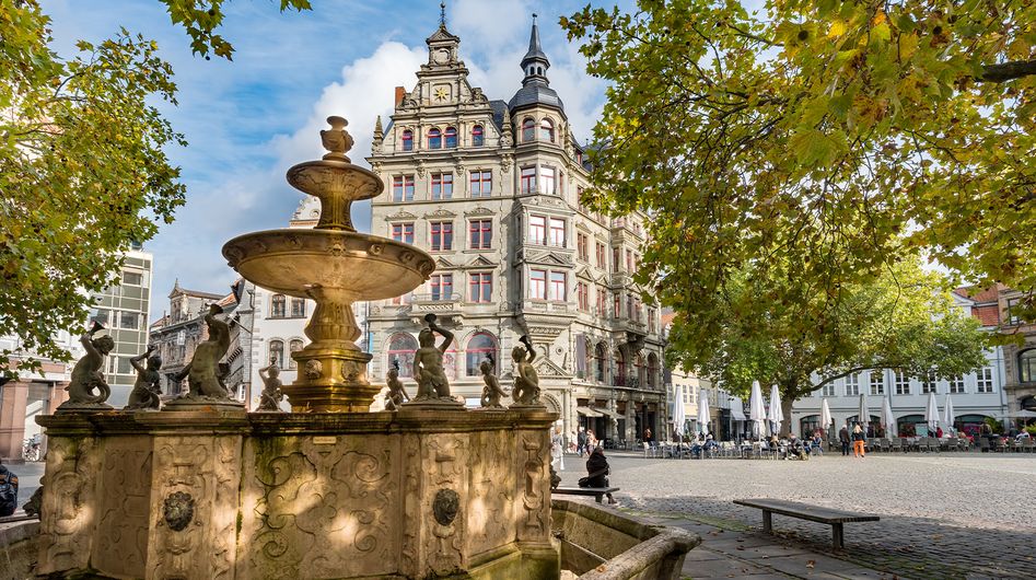 Old fountain under green trees in front of a historic building in Braunschweig