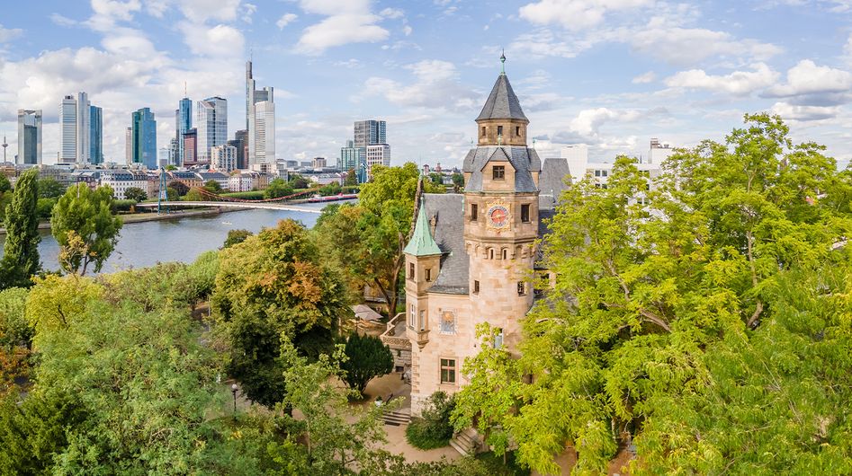 Historisches Gebäude umgeben von grünen Bäumen, im Hintergrund der Main und das Bankenviertel von Frankfurt am Main