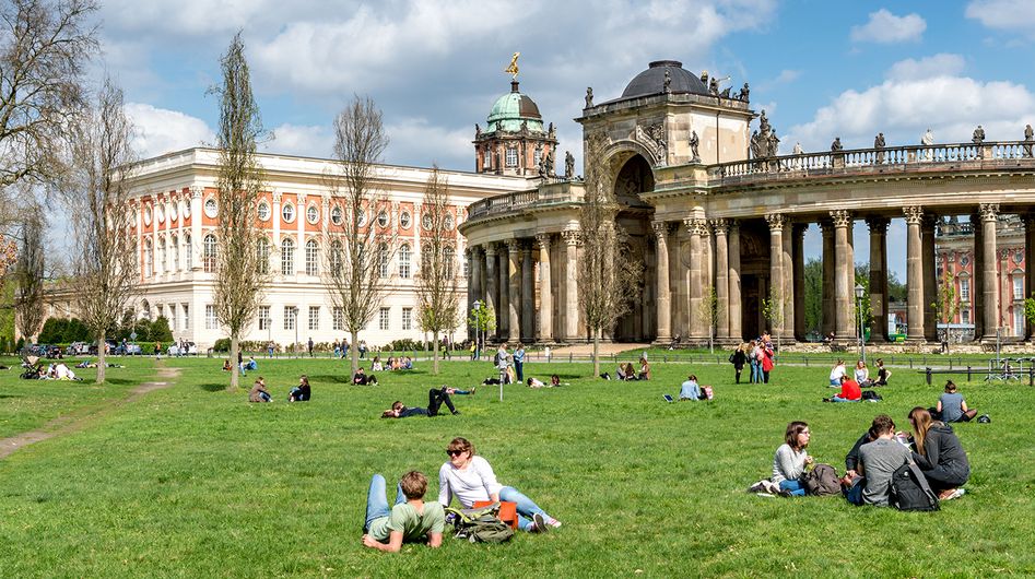 Junge Menschen sitzen gemeinsam auf dem Rasen in Potsdam, im Hintergrund historische Gebäude.