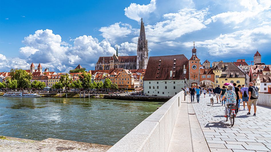 Menschen überqueren ein Brücke, im Hintergrund der Regensburger Dom und historische Gebäude.