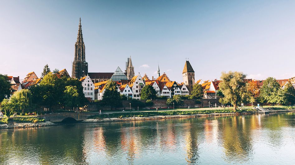 Blick über einen Fluss auf Häuser am Ufer, im Hintergrund das Ulmer Münster