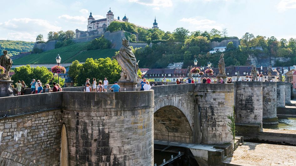 In the foreground the Old Main River Bridge in Würzburg with people, in the background the Marienberg Fortress